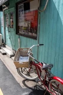 A bright red bicycle with a wicker basket rests against a turquoise wooden wall outside a shop. The shop has a large window displaying various bottles and posters, and a door painted in the same turquoise. The scene is illuminated by natural sunlight, casting shadows on the ground.