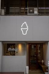 A storefront features a large, illuminated ice cream cone icon above the entrance. The window displays the words 'Soft Daimyo Cream' in stylized lettering. The facade is modern and minimalist, with a neutral color palette. Inside, a soft serve ice cream cone decoration is visible near the door.