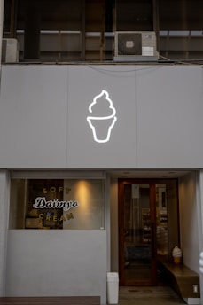 A storefront features a large, illuminated ice cream cone icon above the entrance. The window displays the words 'Soft Daimyo Cream' in stylized lettering. The facade is modern and minimalist, with a neutral color palette. Inside, a soft serve ice cream cone decoration is visible near the door.
