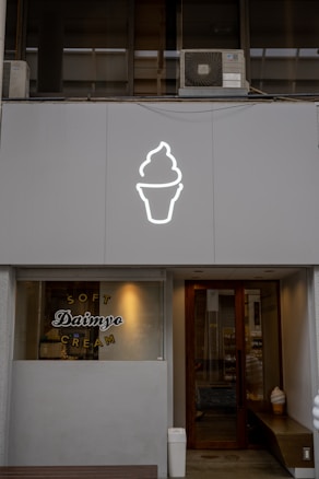 A storefront features a large, illuminated ice cream cone icon above the entrance. The window displays the words 'Soft Daimyo Cream' in stylized lettering. The facade is modern and minimalist, with a neutral color palette. Inside, a soft serve ice cream cone decoration is visible near the door.
