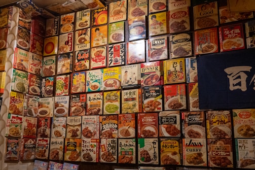 A wall covered with vintage curry packaging boxes, each featuring colorful designs and illustrations of various curry dishes and branding. The boxes create a vibrant collage with diverse fonts and graphics, giving a nostalgic feel. There is a mix of Japanese and English text, suggesting a range of curry products from different eras.