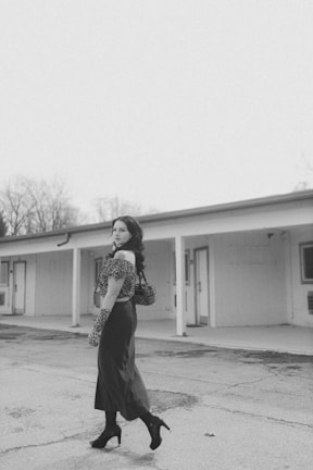 A woman in a vintage-inspired outfit stands in an outdoor corridor of a simple, single-story building. She wears high heels, a long skirt, and a leopard print top and gloves. Her pose is confident and she carries a stylish handbag.