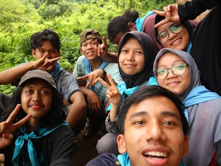 A group of friends is gathered outdoors, smiling and making peace signs for a selfie. They are wearing casual clothing and appear to be enjoying a pleasant day in a lush, green environment.