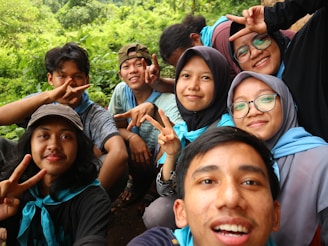 A group of friends is gathered outdoors, smiling and making peace signs for a selfie. They are wearing casual clothing and appear to be enjoying a pleasant day in a lush, green environment.