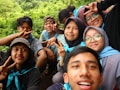 A group of friends is gathered outdoors, smiling and making peace signs for a selfie. They are wearing casual clothing and appear to be enjoying a pleasant day in a lush, green environment.