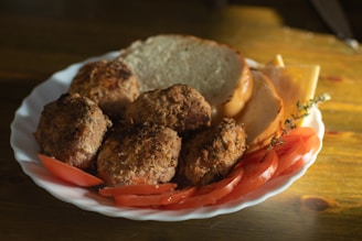 A white plate features several browned meatballs accompanied by slices of fresh tomato, cheese, and a couple of slices of bread. A sprig of thyme is visible, adding a touch of green to the warm tones. The image appears to be shot in natural light, casting soft shadows on the wooden table.