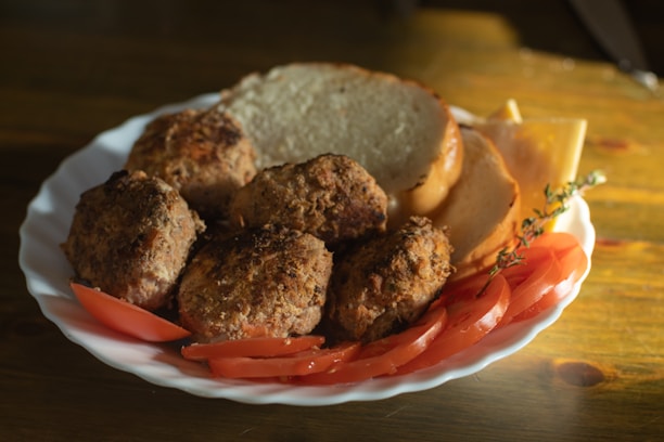 A white plate features several browned meatballs accompanied by slices of fresh tomato, cheese, and a couple of slices of bread. A sprig of thyme is visible, adding a touch of green to the warm tones. The image appears to be shot in natural light, casting soft shadows on the wooden table.