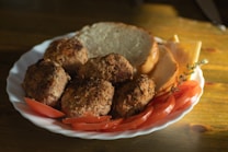 A white plate features several browned meatballs accompanied by slices of fresh tomato, cheese, and a couple of slices of bread. A sprig of thyme is visible, adding a touch of green to the warm tones. The image appears to be shot in natural light, casting soft shadows on the wooden table.