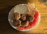A white plate contains four browned meat patties, slices of fresh tomato, two pieces of bread, some slices of cheese, and a sprig of fresh herbs. The food is arranged on a wooden table, and the scene is well-lit, suggesting a natural and warm atmosphere.