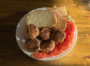 A white plate contains four browned meat patties, slices of fresh tomato, two pieces of bread, some slices of cheese, and a sprig of fresh herbs. The food is arranged on a wooden table, and the scene is well-lit, suggesting a natural and warm atmosphere.