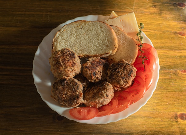 A white plate contains four browned meat patties, slices of fresh tomato, two pieces of bread, some slices of cheese, and a sprig of fresh herbs. The food is arranged on a wooden table, and the scene is well-lit, suggesting a natural and warm atmosphere.