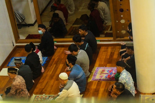 A group of people is seated on prayer mats on a wooden floor, engaged in a religious gathering or ritual. Some participants are wearing hats and face masks. The environment appears to be calm and focused, with a partition or wall depicting decorative cutouts separating them from another area.