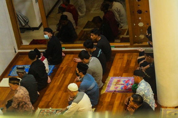 A group of people is seated on prayer mats on a wooden floor, engaged in a religious gathering or ritual. Some participants are wearing hats and face masks. The environment appears to be calm and focused, with a partition or wall depicting decorative cutouts separating them from another area.