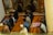 A group of people is seated on prayer mats on a wooden floor, engaged in a religious gathering or ritual. Some participants are wearing hats and face masks. The environment appears to be calm and focused, with a partition or wall depicting decorative cutouts separating them from another area.