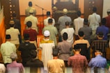 A group of people, all dressed in various types of traditional and modern attire, are seen gathered together, standing in rows, with some bowed in prayer. The setting appears to be indoors, with a wooden podium or stand at the front. A microphone stands ready for use, suggesting a communal or religious gathering.