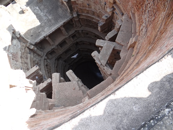 A deep, ancient stepwell constructed from stone and brick with intricate carvings lining the circular walls. Numerous stone steps and platforms lead downwards into darkness, suggesting great depth. Sunlight illuminates parts of the structure, casting shadows and highlighting the detailed architecture.
