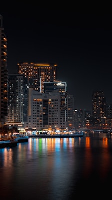 Nighttime city skyline with illuminated buildings reflecting on calm water.