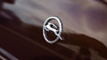 A chrome emblem in the shape of a circle with a horizontal line and a stylized car figure is affixed to a brown metallic surface, likely a vehicle. The surface displays a subtle reflection.