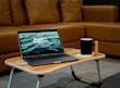 A laptop displaying the text 'Building Websites' sits on a wooden portable table in front of a brown sofa. Next to the laptop, there is a black mug placed on a white coaster. The setting suggests a cozy indoor environment suited for remote work or relaxation.