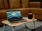 A laptop displaying the text 'Building Websites' sits on a wooden portable table in front of a brown sofa. Next to the laptop, there is a black mug placed on a white coaster. The setting suggests a cozy indoor environment suited for remote work or relaxation.