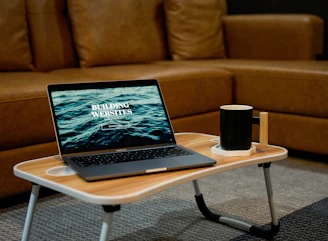 A laptop displaying the text 'Building Websites' sits on a wooden portable table in front of a brown sofa. Next to the laptop, there is a black mug placed on a white coaster. The setting suggests a cozy indoor environment suited for remote work or relaxation.