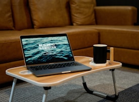 A laptop displaying the text 'Building Websites' sits on a wooden portable table in front of a brown sofa. Next to the laptop, there is a black mug placed on a white coaster. The setting suggests a cozy indoor environment suited for remote work or relaxation.