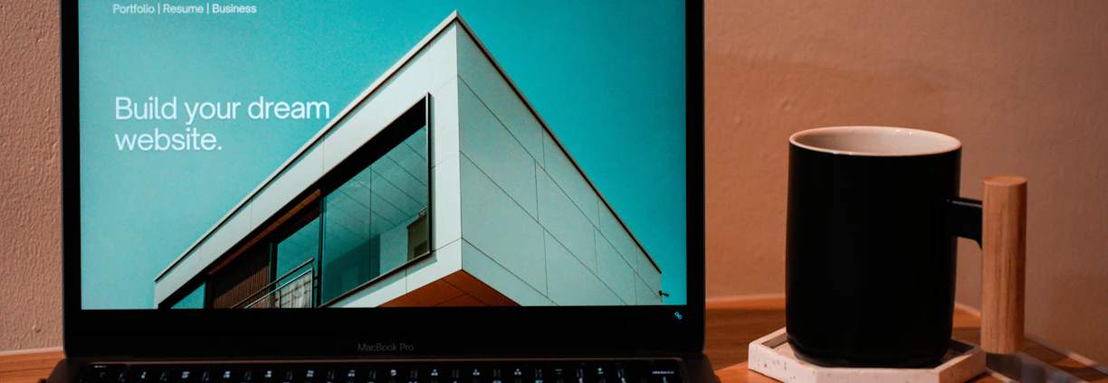 A laptop displaying a website design with the phrase 'Build your dream website.' next to a minimalistic building image. The scene also includes a modern mug and a smartphone placed on a wooden desk.