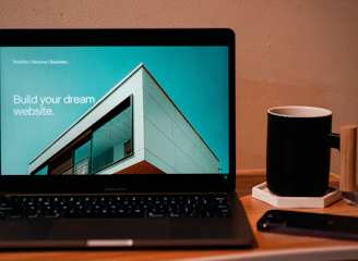 A laptop displaying a website design with the phrase 'Build your dream website.' next to a minimalistic building image. The scene also includes a modern mug and a smartphone placed on a wooden desk.