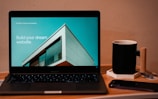 A laptop displaying a website design with the phrase 'Build your dream website.' next to a minimalistic building image. The scene also includes a modern mug and a smartphone placed on a wooden desk.