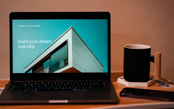 A laptop displaying a website design with the phrase 'Build your dream website.' next to a minimalistic building image. The scene also includes a modern mug and a smartphone placed on a wooden desk.