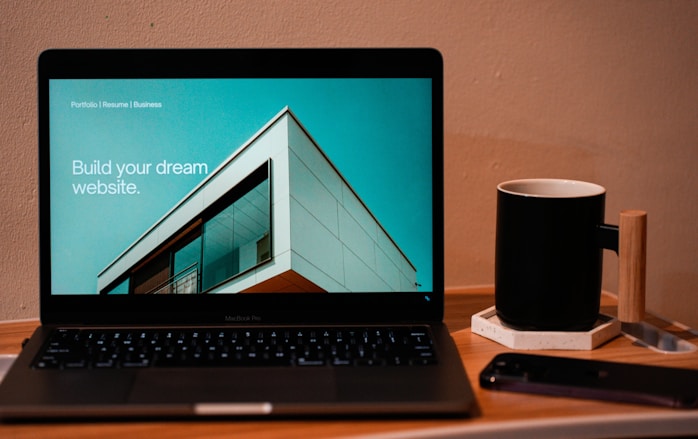 A laptop displaying a website design with the phrase 'Build your dream website.' next to a minimalistic building image. The scene also includes a modern mug and a smartphone placed on a wooden desk.