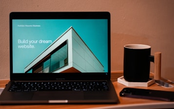 A laptop displaying a website design with the phrase 'Build your dream website.' next to a minimalistic building image. The scene also includes a modern mug and a smartphone placed on a wooden desk.