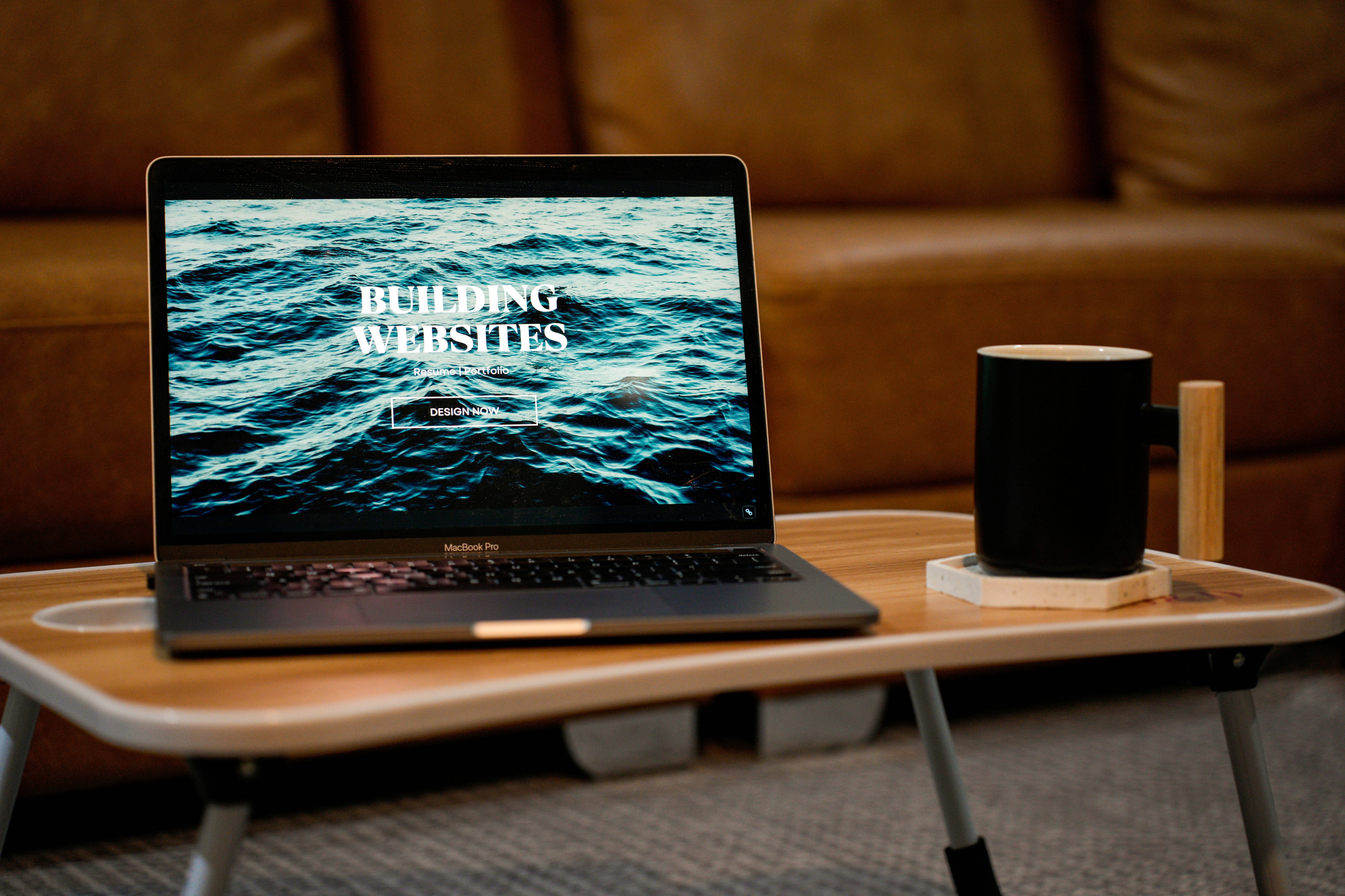 A laptop, MacBook, on top of a portable table with coffee tea mug on the side.