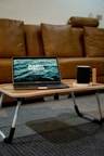 A laptop displaying the words 'Building Websites' rests on a small wooden table with black and white legs. Next to the laptop is a black mug placed on a coaster. The setting includes a brown leather sofa in the background and a patterned carpeted floor.