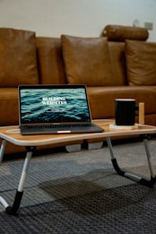 A laptop displaying the words 'Building Websites' rests on a small wooden table with black and white legs. Next to the laptop is a black mug placed on a coaster. The setting includes a brown leather sofa in the background and a patterned carpeted floor.