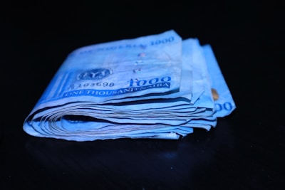A stack of folded blue banknotes with a denomination of one thousand naira is placed on a dark surface.