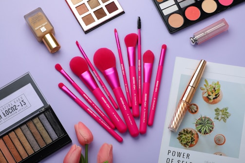 A flat lay of various makeup items arranged on a light purple surface. The central focus is on a set of vibrant pink makeup brushes, surrounded by eyeshadow palettes, a bottle of foundation, a lipstick, and mascara. A couple of pink tulips add a fresh touch, while a magazine featuring green succulents is also partially visible.