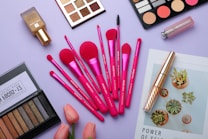 A flat lay of various makeup items arranged on a light purple surface. The central focus is on a set of vibrant pink makeup brushes, surrounded by eyeshadow palettes, a bottle of foundation, a lipstick, and mascara. A couple of pink tulips add a fresh touch, while a magazine featuring green succulents is also partially visible.
