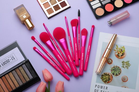 A flat lay of various makeup items arranged on a light purple surface. The central focus is on a set of vibrant pink makeup brushes, surrounded by eyeshadow palettes, a bottle of foundation, a lipstick, and mascara. A couple of pink tulips add a fresh touch, while a magazine featuring green succulents is also partially visible.