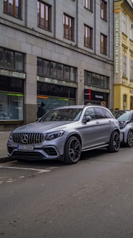 A silver luxury SUV is parked on a city street next to a sidewalk. The vehicle is positioned beside a building with large windows and a sign indicating the presence of a cinema, shopping, and garden. The street is relatively empty, with only a few pedestrians visible.