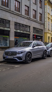 A silver luxury SUV is parked on a city street next to a sidewalk. The vehicle is positioned beside a building with large windows and a sign indicating the presence of a cinema, shopping, and garden. The street is relatively empty, with only a few pedestrians visible.
