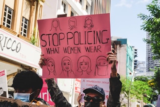 A person holds a pink protest sign in a street setting. The sign features illustrations of various women and a message advocating for women's rights, specifically addressing the policing of women's clothing. Several individuals are gathered around, some wearing masks, and buildings and trees are visible in the background.