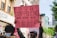A person holds a pink protest sign in a street setting. The sign features illustrations of various women and a message advocating for women's rights, specifically addressing the policing of women's clothing. Several individuals are gathered around, some wearing masks, and buildings and trees are visible in the background.
