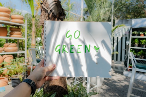 A hand holding a spiral notebook that reads 'GO GREEN' in bright green letters. The background features a garden setting with several terracotta pots, lush green plants, and palm leaves. Sunlight casts shadows on the notebook, adding to the bright and cheerful ambiance.