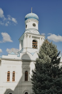 A white church with architectural features such as arched windows and decorative details on the façade. The church has a prominent light blue dome adorned with a cross at the top and a clock below the dome. A tall evergreen tree partially obscures the view of the building's lower section, while a clear blue sky with a few scattered clouds forms the background.