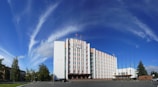A large, multi-story government building with a flag on top and a clear blue sky with wispy clouds. The building is surrounded by trees and has a paved area with flagpoles in front.