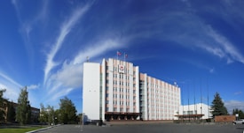A large, multi-story government building with a flag on top and a clear blue sky with wispy clouds. The building is surrounded by trees and has a paved area with flagpoles in front.