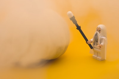 A LEGO figure with long gray hair and beard, dressed in white robes, is holding a staff and facing a large, hazy, and indistinct object. The background is a solid yellow color, adding to the warm tone of the image.