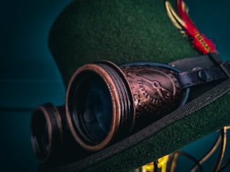 A close-up image of a green top hat adorned with vintage-style brown goggles. The hat also features a red and yellow feather as decoration.