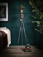 A steampunk-inspired lamp with a metal base and vintage light bulb is topped with a green bowler hat. The lamp stands against a dark teal paneled wall next to a leafy plant and a framed print. On the wooden floor beside the lamp, a stack of books supports a matching green cup and saucer.